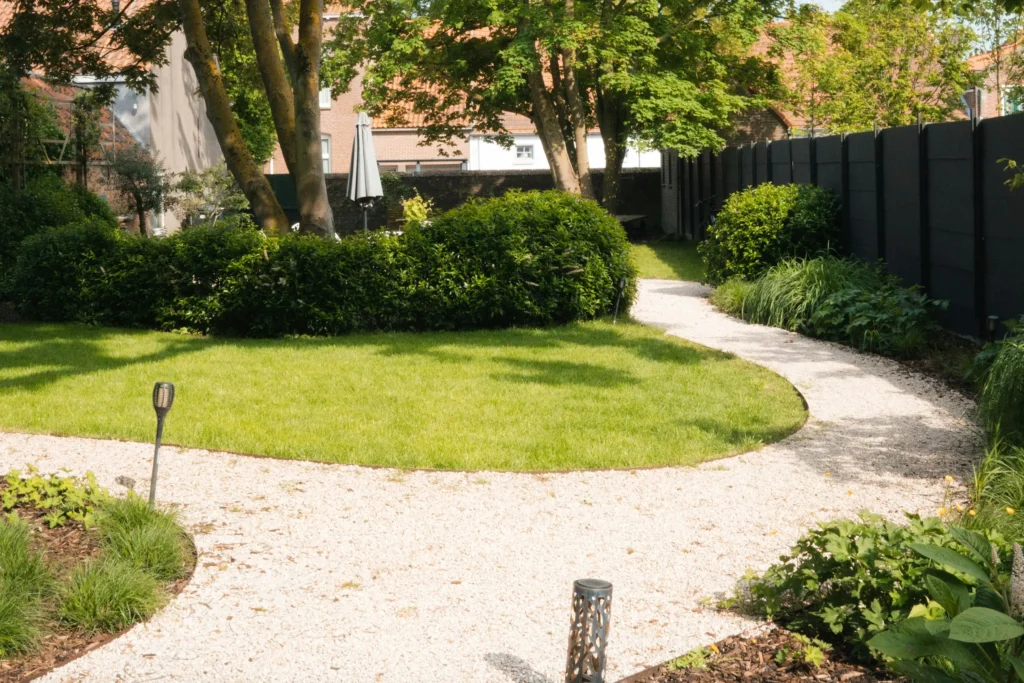 Curved white gravel garden path surrounding a green lawn with mature trees, clipped shrubs, ornamental grasses, and dark privacy fencing.