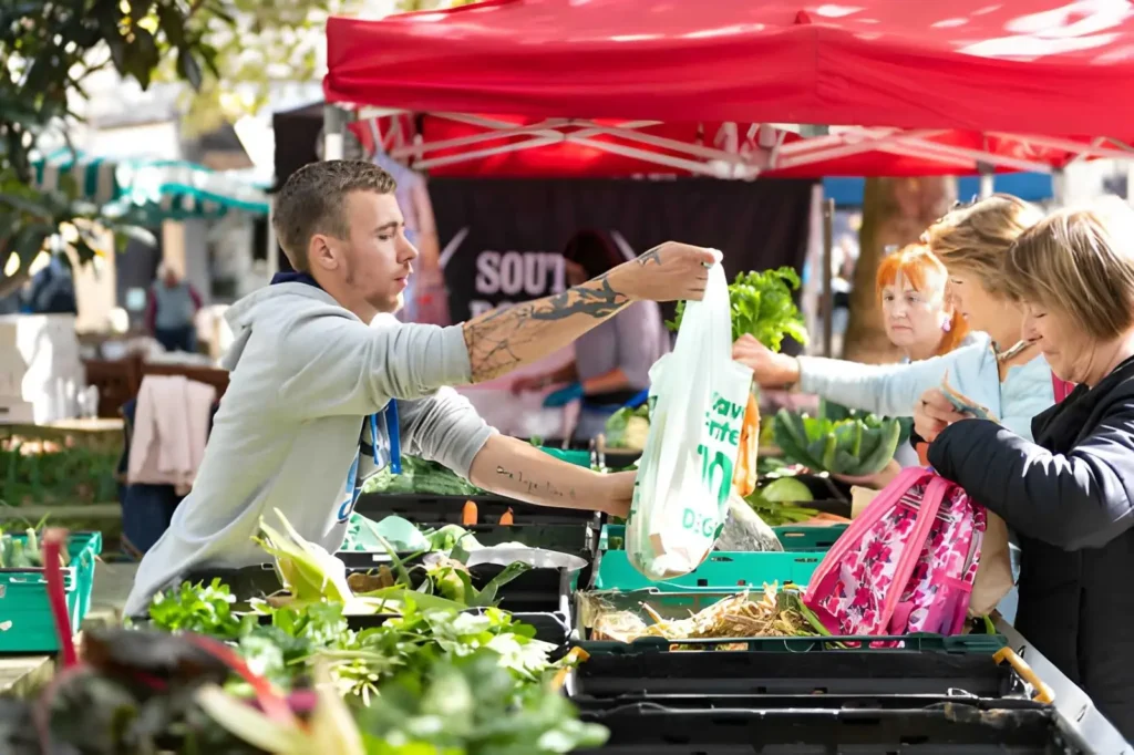 Farmers market assistant handing a bag of fresh vegetables to customers at an outdoor produce stand, illustrating a homesteading job that does not require owning a large farm.