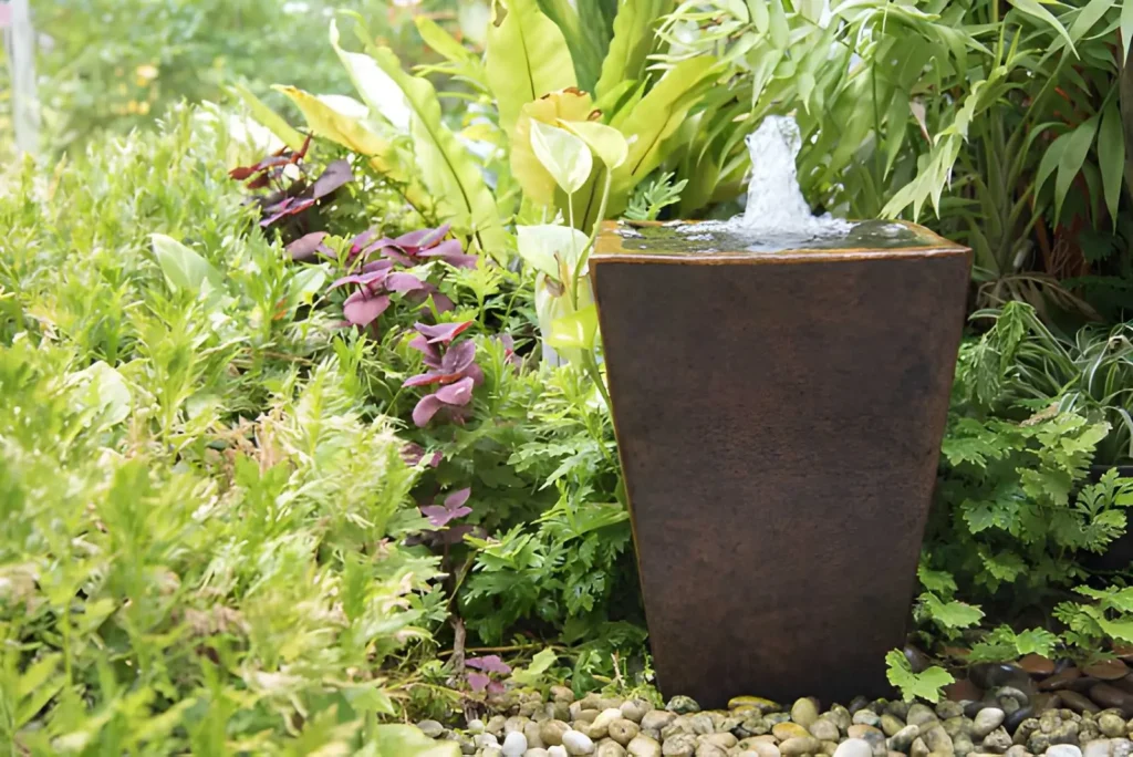 Modern dark container fountain with bubbling water surrounded by lush green plants and decorative river stones in a small garden.