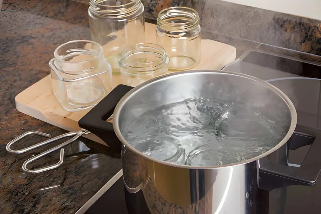 Empty glass canning jars being preheated in a pot of hot water beside a jar lifter and additional clean jars on a kitchen counter.