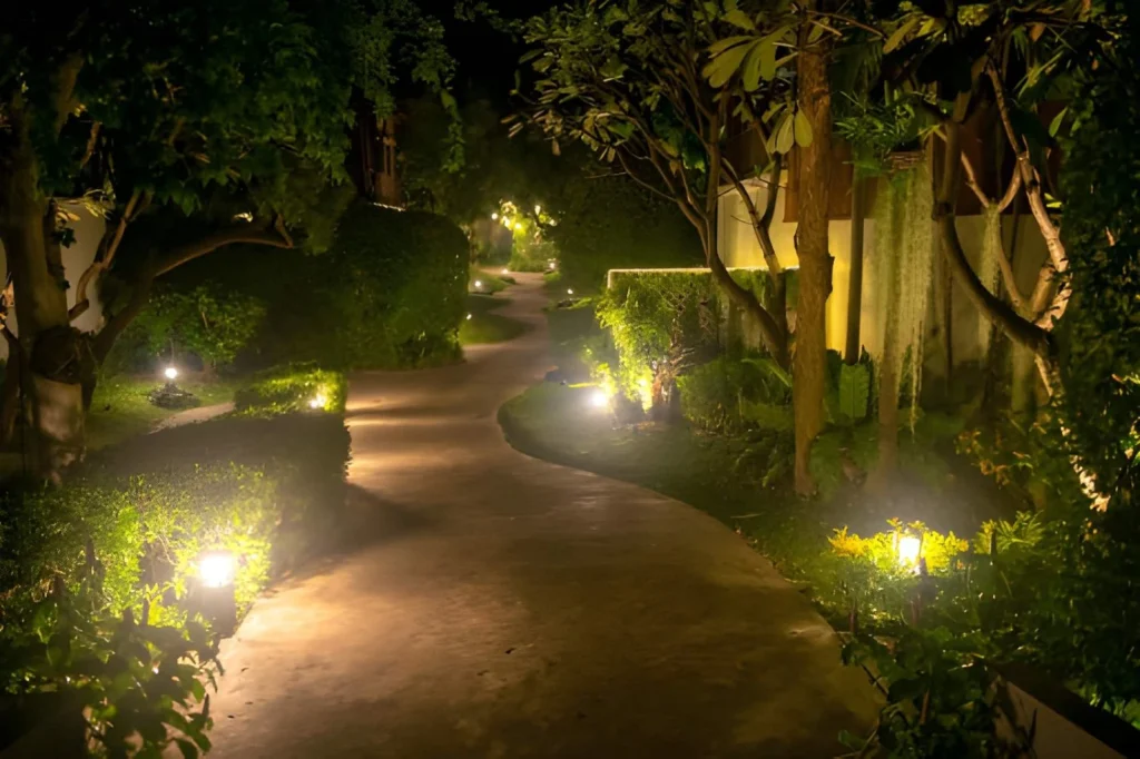 Small garden design with warm landscape lighting illuminating a curved pathway bordered by trees and neatly trimmed greenery.