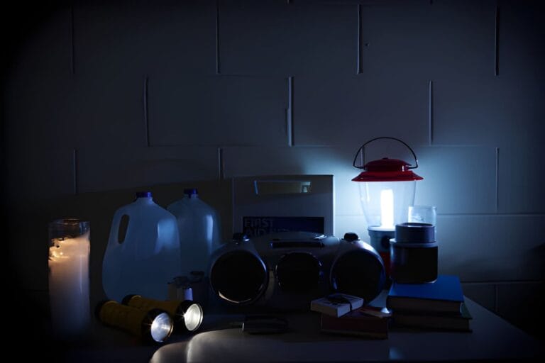 Power outage preparedness supplies on a homestead table with lanterns, water containers, canned food, radio, first aid kit, and blankets.