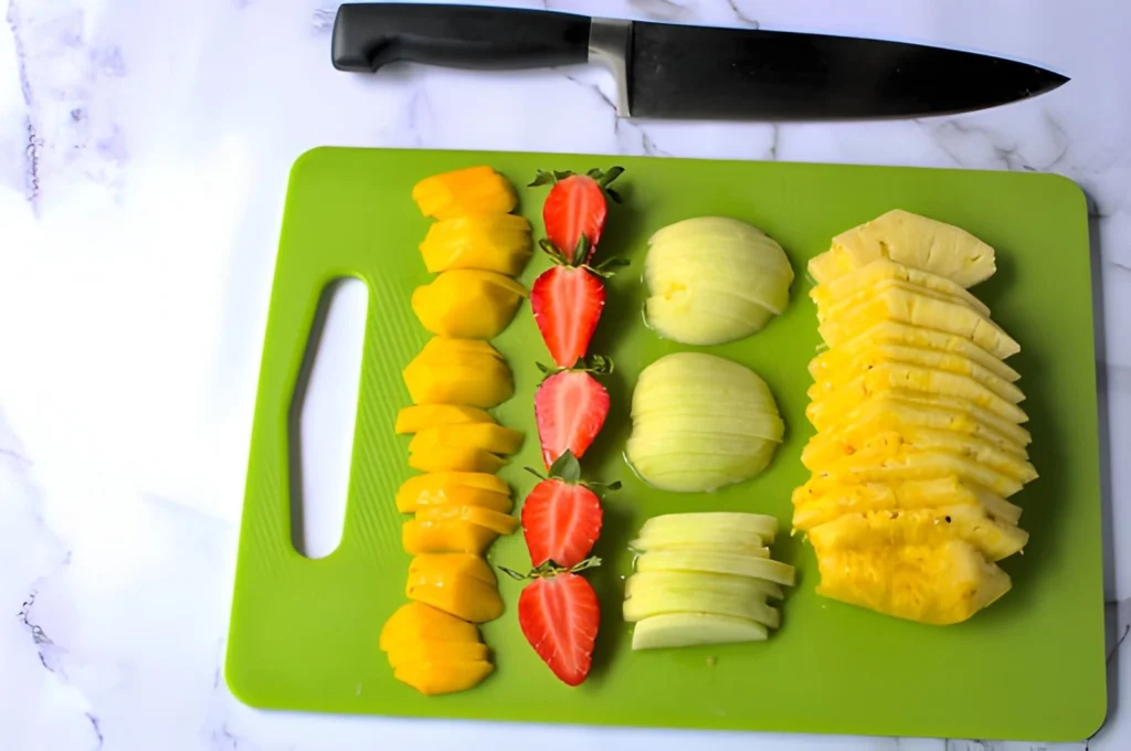 Evenly sliced mango, strawberries, apples, and pineapple arranged on a cutting board before dehydrating for beginner food preservation.