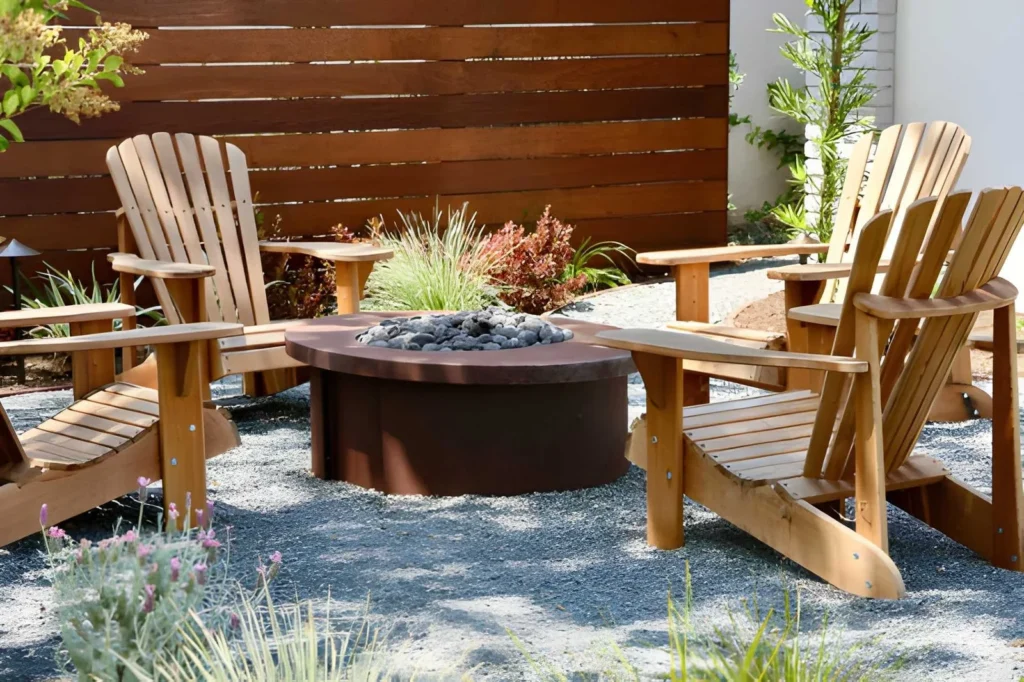 Cozy small backyard fire pit area with wooden Adirondack chairs, gravel ground cover, privacy fencing, and surrounding ornamental plants.